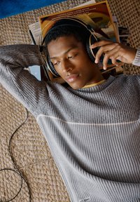 Siv šeširasti pulover z žametenim vzorcem, ki ima bel trak. Črne slušalke z obokom in zlatim poudarkom. Ležijo na tkani preprogi.