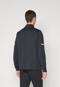 Svart langermet polo med krage, som har en neonfarge på venstre erm. Laget av mykt, teksturert stoff for komfort.