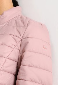 Chaqueta acolchada de color rosa claro con un cuello alto. Presenta costuras verticales, textura suave y un pequeño parche con el logo en la manga.