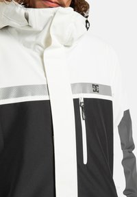 Giacca impermeabile bianca e nera con accenti grigi, caratterizzata da un colletto alto, zip frontale, tasca sul petto e strisce riflettenti sul petto.