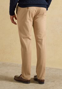 Homme portant un pantalon beige, des mocassins marron foncé et un pull en maille torsadée bleu marine, debout sur un sol texturé devant un mur beige.