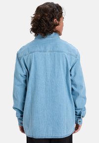 Lichtblauw denim overhemd met lange mouwen, knoopmanchetten en een klassieke kraag. Bevat een gestikte naaddetail over de achterkant.