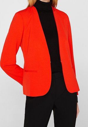 Veste blazer rouge vif cintrée portée sur un col roulé noir et un pantalon noir, visible des épaules aux hanches.