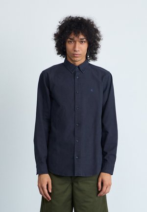 Chemise boutonnée bleu marine en coton avec col classique, manches longues et un petit logo sur la gauche de la poitrine. Texture lisse.
