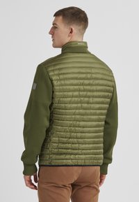 Olivengrüne Jacke mit einer gepolsterten, gesteppten Textur auf dem Rücken, weichen Strickstoffärmeln, hohem Kragen und gerippten Bündchen.