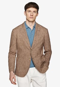 Uomo che indossa un blazer marrone testurizzato sopra una polo blu e pantaloni bianchi, in piedi con una mano in tasca contro uno sfondo semplice.