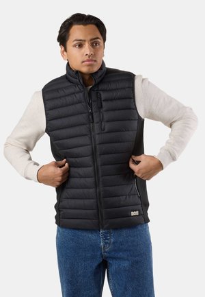 Jeune homme fermant une veste matelassée noire sans manches par-dessus une chemise beige clair à manches longues, portant un jean bleu.