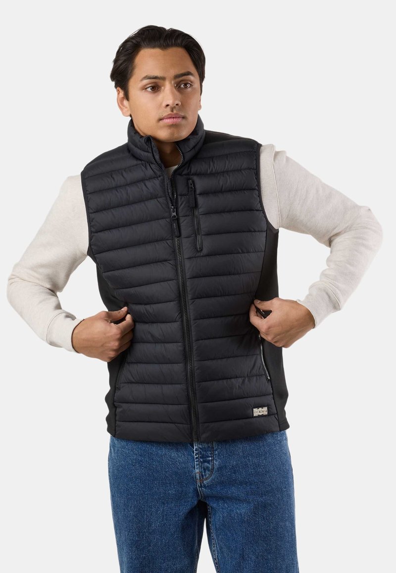 Giovane che chiude la cerniera di un gilet imbottito nero senza maniche sopra una camicia beige chiaro a maniche lunghe, indossando jeans blu.