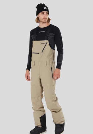 Beige waterdichte overall met verstelbare banden, voorzakken met rits, verstevigde knieën en zwarte accenten op de onderbenen. Zwarte beanie gedragen.