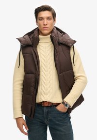 Braune, puffy Weste mit hohem Kragen, über einem cremefarbenen Norwegerpullover getragen. Kombiniert mit dunkelblauen Jeans und einer Uhr.