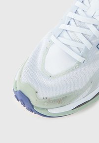 Lys blå atletisk sko med teksturert mesh-overdel, hvit mellomsåle, gjennomsiktige mintgrønne detaljer og lilla yttersåle.