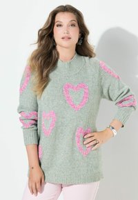 Kobieta ubrana w jasnoszary sweter z dzianiny z różowymi sercami, jasnoróżowe spodnie i srebrne bransoletki, stojąca z jedną ręką na biodrze.