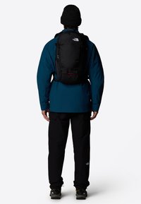 Mochila negra con correas ajustables, que presenta un acento rojo, llevada sobre una chaqueta azul y pantalones negros, ubicada sobre un fondo blanco.