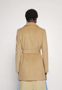 Camelwollen jas met een getailleerde pasvorm, geknipt kraag en een riem in de taille. Soepele textuur en een middenlengte snit.