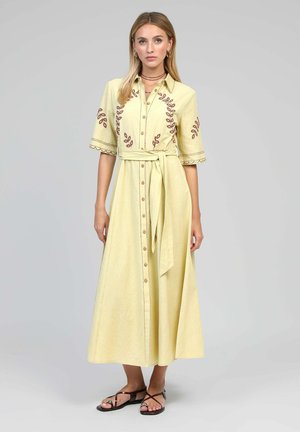 Femme portant une robe midi boutonnée jaune clair avec une broderie de feuilles brunes, une ceinture nouée à la taille, des manches au coude et des sandales noires à lanières.