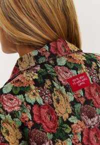 Kwiatowy materiał żakardowy w czerni z wielokolorowymi różami. Czerwona metka z białym tekstem wszyta w kołnierz, brzmiąca "ZA CHWILĘ STWORZYSZ MAGIĘ."