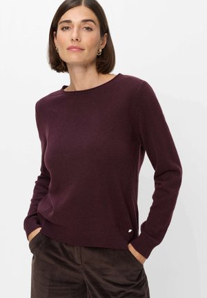 Donna con capelli corti castani indossa un maglione viola scuro a maniche lunghe e pantaloni marroni, in piedi con le mani nelle tasche, rivolta in avanti.