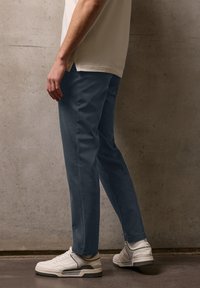 Mężczyzna ubrany w niebieskie spodnie, białe trampki i kremową koszulę stojący przy szarej betonowej ścianie.