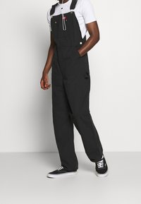 Salopette nera realizzata in un tessuto robusto, con spalline regolabili, tasche laterali e una tasca frontale. Abbinata a una t-shirt bianca e sneakers nere.