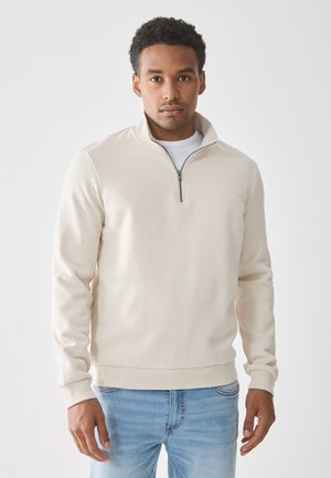 Sudadera beige con cremallera, de manga larga, cuello alto y puños acanalados. Tela texturizada; combinada con jeans de mezclilla azul claro.