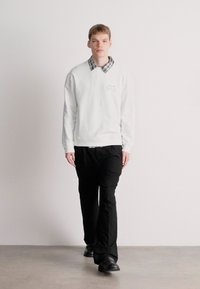 Λευκή μπλούζα με στρογγυλή λαιμόκοψη, μαύρο παντελόνι και μαύρα παπούτσια. Η μπλούζα διαθέτει καρό γιακά και διακριτική λεπτομέρεια κειμένου.