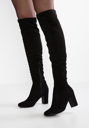 Zwarte suède kniehoge laarzen met blokhakken en ritssluitingen aan de zijkant, gedragen met doorschijnende zwarte panty's op een witte achtergrond.