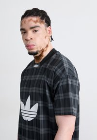 Juoda ir pilka languota trumpomis rankovėmis marškinėliai su dideliu baltu Adidas trefoil logotipu priekyje, su apvaliu kaklu ir lygiu paviršiumi.