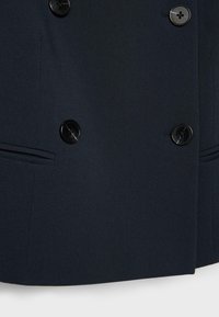 Manteau bleu marine foncé à double boutonnage avec boutons noirs à quatre trous et poches passepoilées horizontales de chaque côté.
