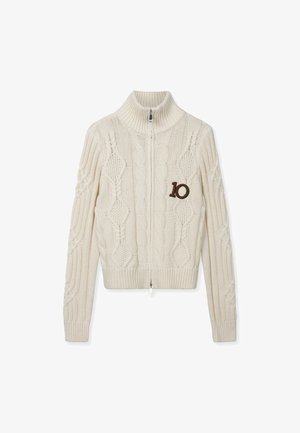 Roomkleurige kabelgebreide trui met ritssluiting, hoge kraag en een donkerbruine patch met het nummer "10" op de linkerborst.