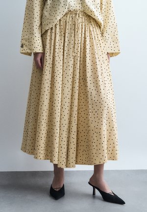 Μπεζ μίντι φούστα με πουά και σούρες στη μέση, σετ με μακρυμάνικο τοπ στο ίδιο σχέδιο και μαύρα μυτερά mules με τακούνι.