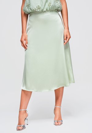 Mujer con falda midi de satén verde claro y sandalias plateadas de tacón alto, de pie contra un fondo sencillo.
