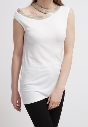 Nainen pukeutuneena valkoiseen hihattomaan toppiin, jossa on epäsymmetrinen kaula-aukko ja kultainen reunus, yhdistettynä mustiin housuihin.