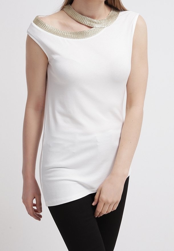 Femme portant un haut blanc sans manches avec un décolleté asymétrique orné d'un liseré doré, associé à un pantalon noir.