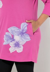 Rosa bomuldstunika med trekvartlange ærmer, der har et stort lyseblåt blomsterprint og en sidelomme for funktionalitet.