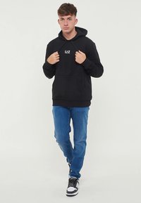 Sweatshirt preto com capuz de cordão e bolso frontal, com logo branco no peito, combinado com jeans azul e ténis pretos.