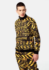 Mustard ja musta värvi lukuga jakk, millel on "QUARANTINE" tekst, bioohtlikkuse sümbolid ja triibuline muster. Valmistatud siledast kangast, millel on soonilised varrukakummid.