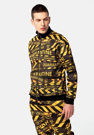 Sort og gul lynlåsjakke med teksten "QUARANTINE", biohazard-symboler og et stribet mønster. Lavet af glat stof med ribbede manchetter.