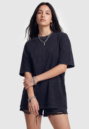 Ung kvinde med langt mørkt hår iført sort pletskudt oversize T-shirt, sorte slidte shorts, sølvkæde halskæde, stjerneformede øreringe og armbånd.