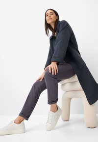 Grå uldfrakke med rund krave, kombineret med tætsiddende mørke bukser og hvide sneakers. Baggrunden er ensfarvet hvid; modellen poser siddende på en lys stol.