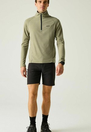 Mężczyzna ubrany w oliwkowozielony longsleeve z zamkiem przy szyi, czarne szorty, czarne skarpetki, czarne buty do biegania i czarny zegarek na nadgarstku, stojący na jasnym tle.