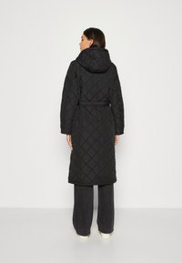 Cappotto lungo trapuntato nero con cappuccio, cintura in vita e maniche oversize. Motivo a diamante testurizzato su tutta la superficie, abbinato a pantaloni scuri.