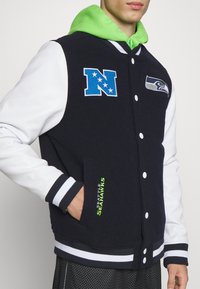 Marineblau und weiße Collegejacke mit grünem Kapuzenaufsatz, versehen mit Seattle Seahawks-Logos und gesticktem Text, entworfen mit gestreiften Rippbündchen.