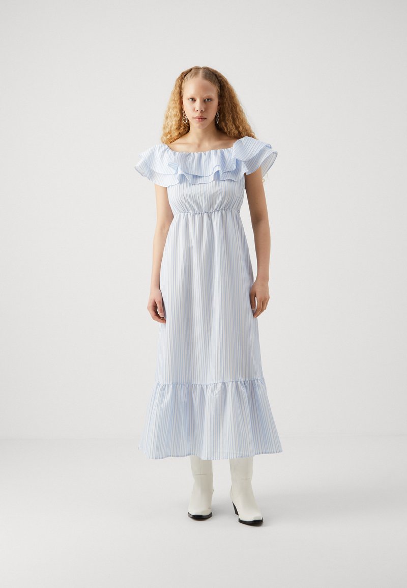 Jasnoniebieska i biała w paski, maxi sukienka z odkrytymi ramionami, z falbaniastym dekoltem i warstwowym dołem. Modelka ma na sobie białe botki na obcasie.