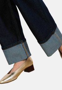 Tmavě modré široké kalhoty z denimu se světle modrým manžetovým lemem a zlatými kovovými plochými botami na dřevěném podpatku, které ukazují kontrastní textury a barvy.