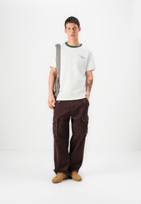 Camiseta blanca con acentos verdes en el cuello, combinada con pantalones cargo marrón oscuro con bolsillos laterales y un corte holgado. Zapatillas color marrón claro.