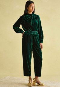 Grüne Samtbluse mit Rüschendetails, kombiniert mit passender Hose. Goldene Absätze runden das Outfit ab. Weicher Stoff, lockere Passform.