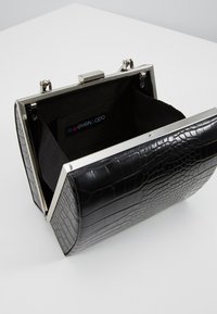 Musta kuvioitu suorakaiteen muotoinen clutch-laukku hopeisella kehyksellä ja lukolla, avoinna näyttäen tyhjän sisätilan ja mustan kangasvuorin, jossa on merkkilappu.