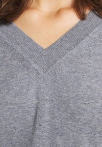 Femme portant un pull en maille gris à col en V, montré des épaules à la poitrine, avec la clavicule visible et des mèches de cheveux bruns.
