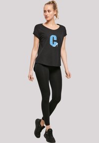 Černé tričko s krátkým rukávem a modrým stylizovaným designem písmene "C", které je spojeno s černými legínami a černými sportovními botami.