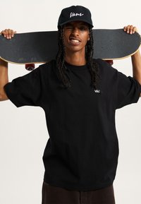 Schwarzes T-Shirt mit gesticktem "Vans"-Logo, kombiniert mit einer schwarzen Mütze und einem Skateboard, das über die Schulter gehalten wird und eine strukturierte Grifffläche aufweist.